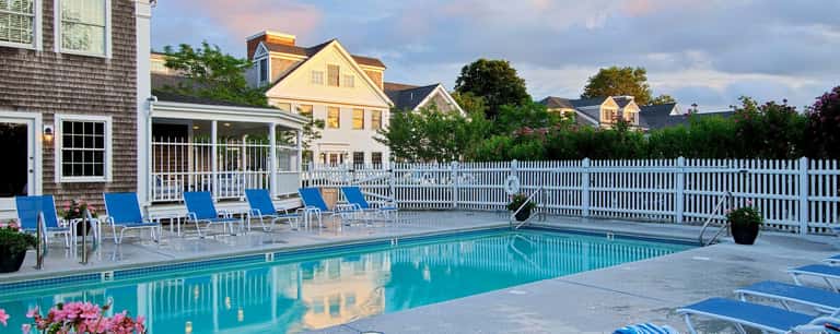 Residential pool area with white fence, blue loungers, and yellow houses in background