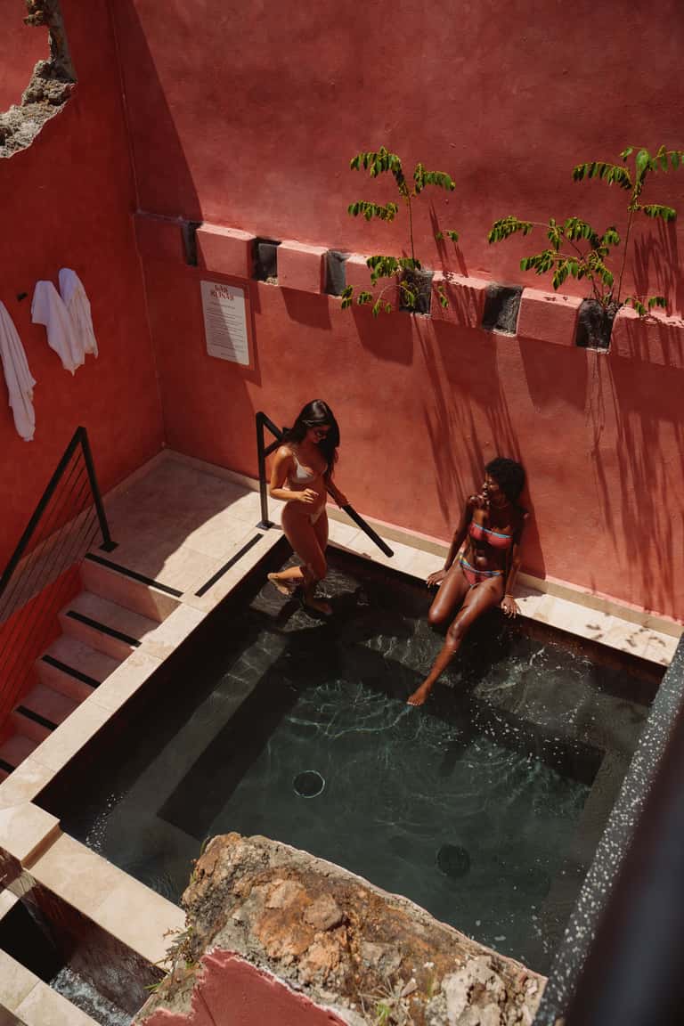 Two women in small dark mineral pool with red walls, plants, and white towels in sunny courtyard