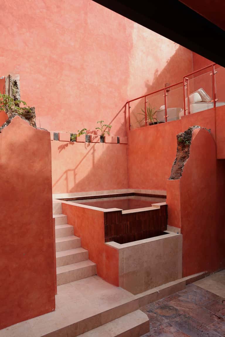 Sunlit thermal pool with wooden accents and stepping stones in a multi-level red spa courtyard
