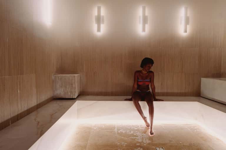 Woman in bikini sitting on edge of illuminated thermal pool with textured beige walls and modern lighting