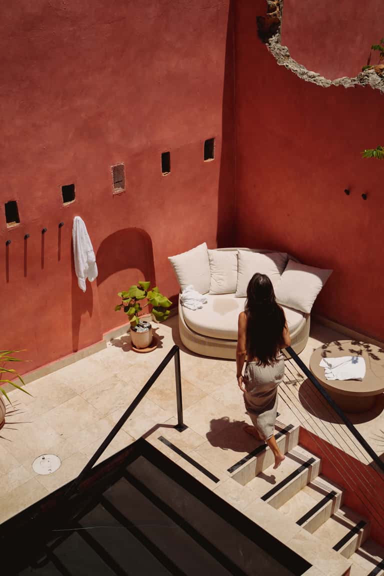 Woman walking down modern steps in terracotta courtyard with cream lounge chair, plants, and minimalist furniture