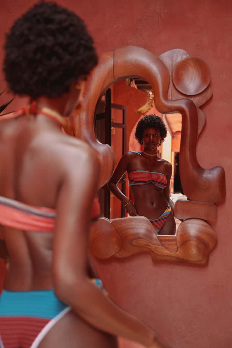 Woman in striped bikini looking in ornate wooden mirror against red wall