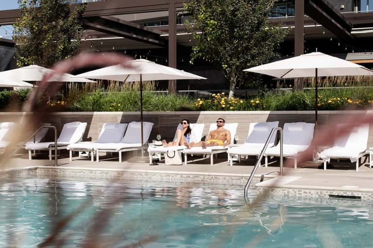 Couple lounging by pool under white umbrellas with landscaped garden and wooden pergola overhead