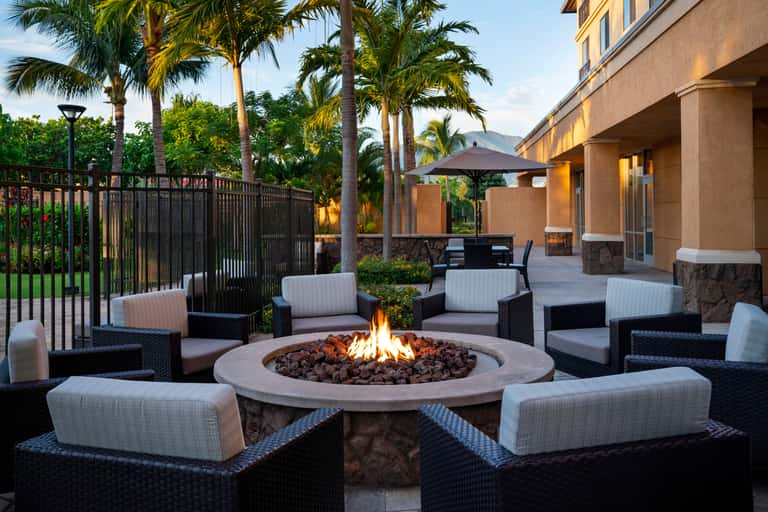 Outdoor fire pit seating area at dusk with black wicker chairs, cushions, palm trees, and illuminated building behind