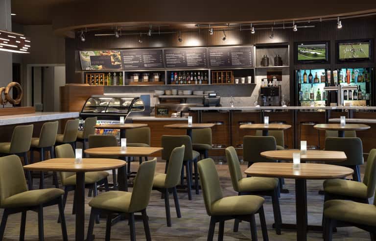 Contemporary cafe or bistro with wooden tables, green upholstered chairs, dark wood counter, shelving, and sports screens