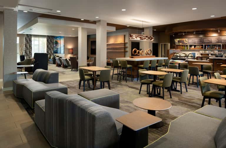 Modern hotel lobby with striped seating, dining tables, bar area, and lounge spaces with warm lighting and contemporary decor