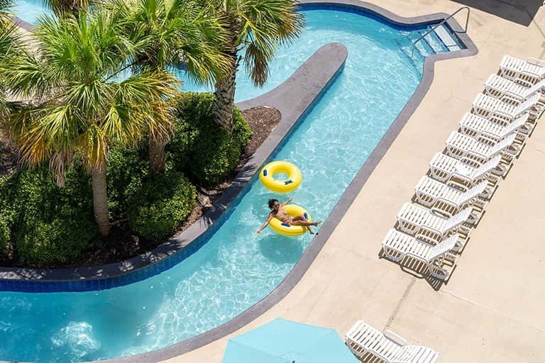 Aerial view of resort lazy river pool with yellow floats, palm trees, and lounge chairs on sunny day