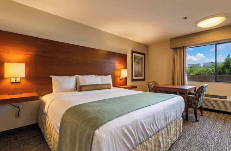 Upscale hotel bedroom with queen bed, wood headboard, warm lighting, desk area, and mountain views through large windows