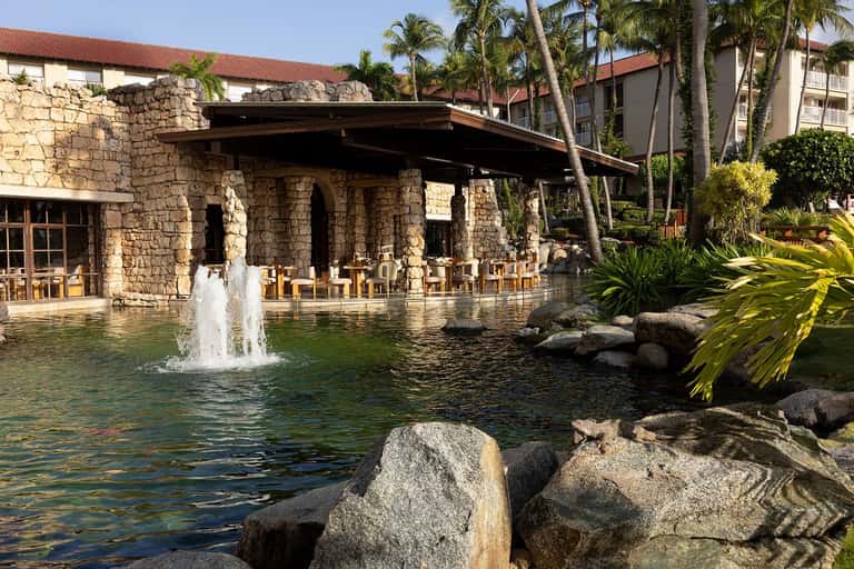 Stone restaurant pavilion with fountain pond, palm trees, and wooden dining chairs overlooking water