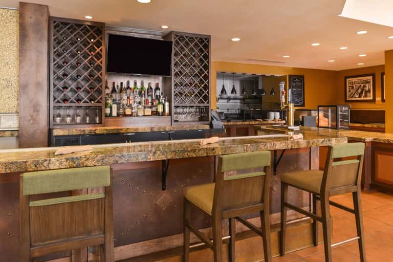 Hotel bar and lounge with granite countertop, wooden bar stools, wine display, amber walls, and full-service bar setup