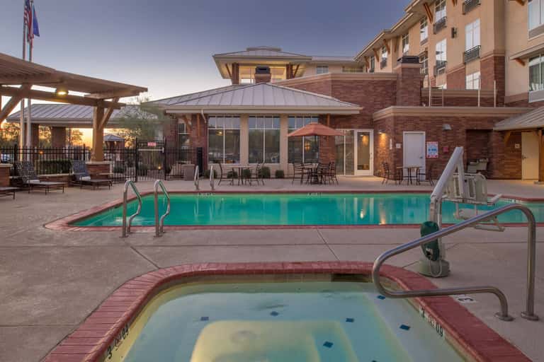 Resort pool and hot tub at dusk with loungers, brick building, and covered patio areas