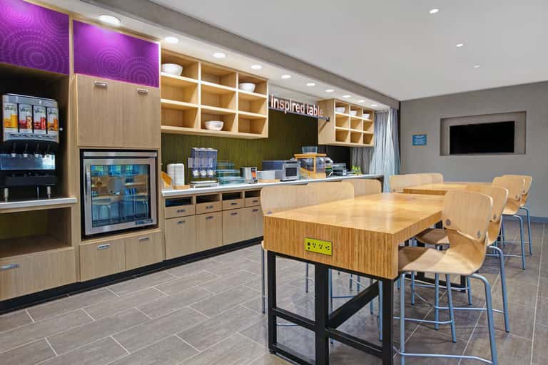 Modern hotel breakfast bar with wood cabinetry, purple accents, seating, and beverage station