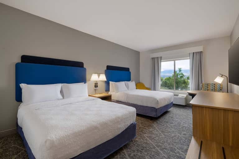 Modern hotel room with two queen beds, blue headboards, white bedding, and panoramic window overlooking mountains