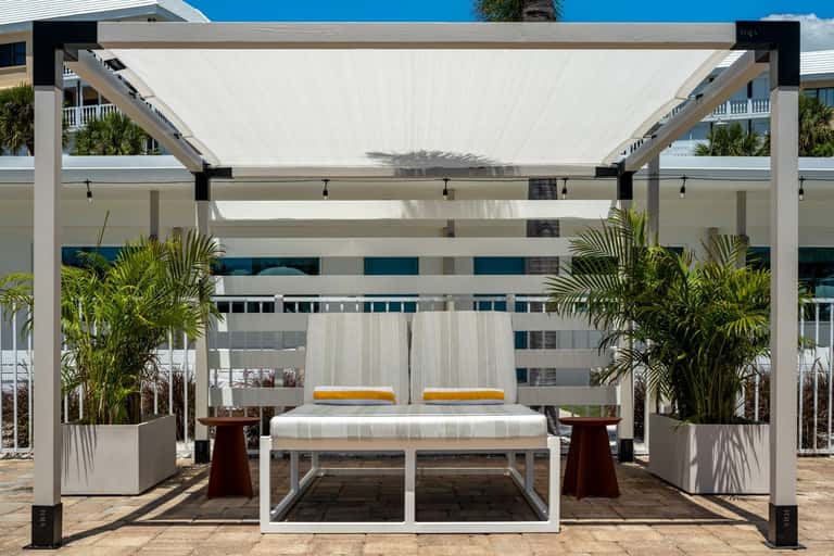 Modern white pergola lounge structure with retractable shade, yellow cushions, and palm planters by the pool