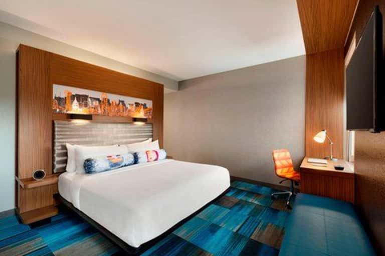 Modern hotel bedroom with wood-paneled headboard, white bedding, blue plaid carpet, and contemporary lighting
