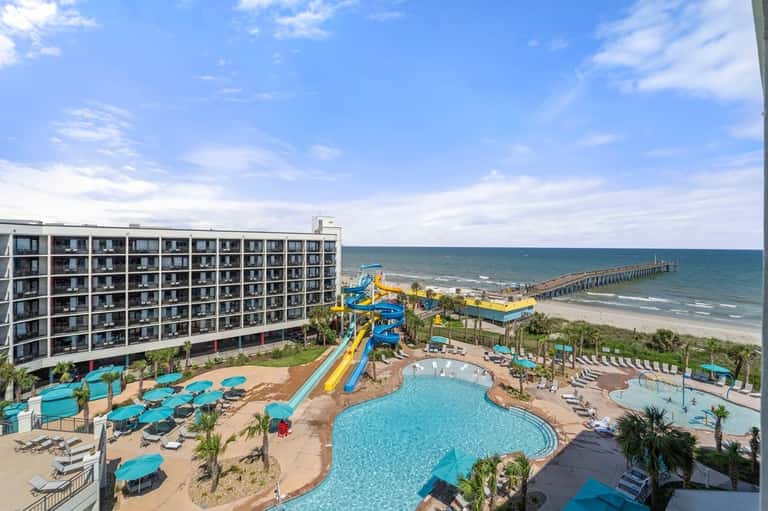 Beachfront resort with pool, water slides, blue umbrellas, palm trees, and ocean view with fishing pier
