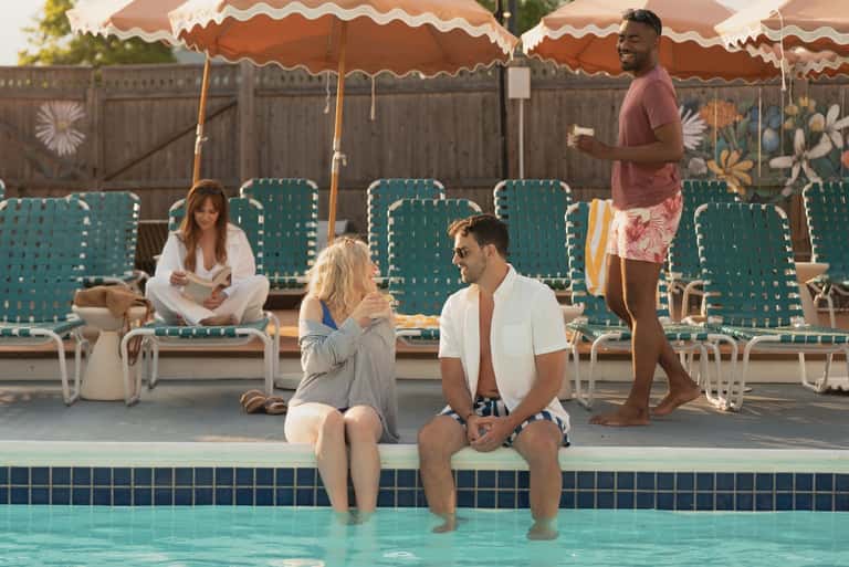 Guests sitting by pool edge under peach umbrellas with teal lounge chairs in background