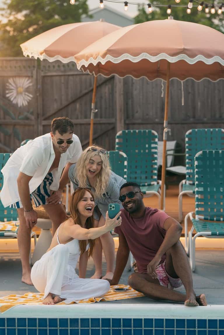Group of four people taking selfie poolside under peach and white scalloped umbrellas
