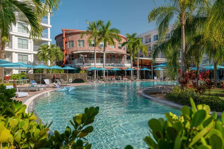 Curved lagoon pool with turquoise water, red brick building, blue umbrellas, and surrounding palms