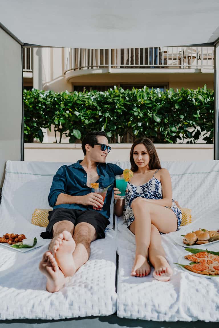 Couple relaxing on white daybed with drinks and food, tropical plants and balconies visible