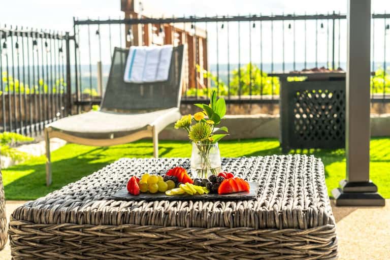 Woven outdoor table with fresh fruit platter and flowers, lounge chairs visible on lawn behind railing