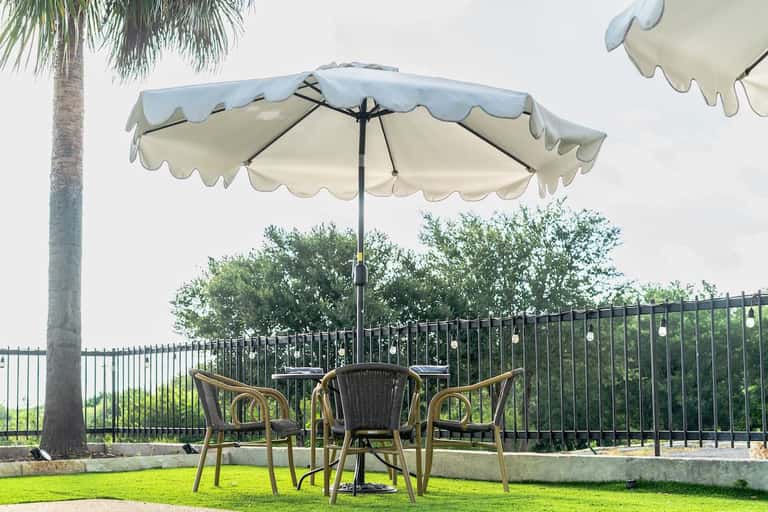 Elegant round table with umbrella, wicker chairs, and manicured lawn overlooking fenced landscape
