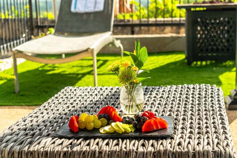 Woven outdoor coffee table with fresh fruit platter and yellow flowers in vase, lounge area behind
