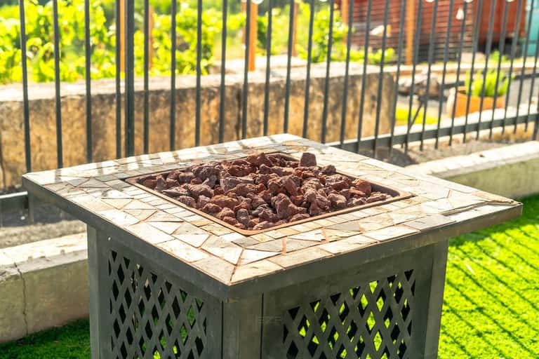 Square fire pit with stone surround on manicured lawn beside black metal railing