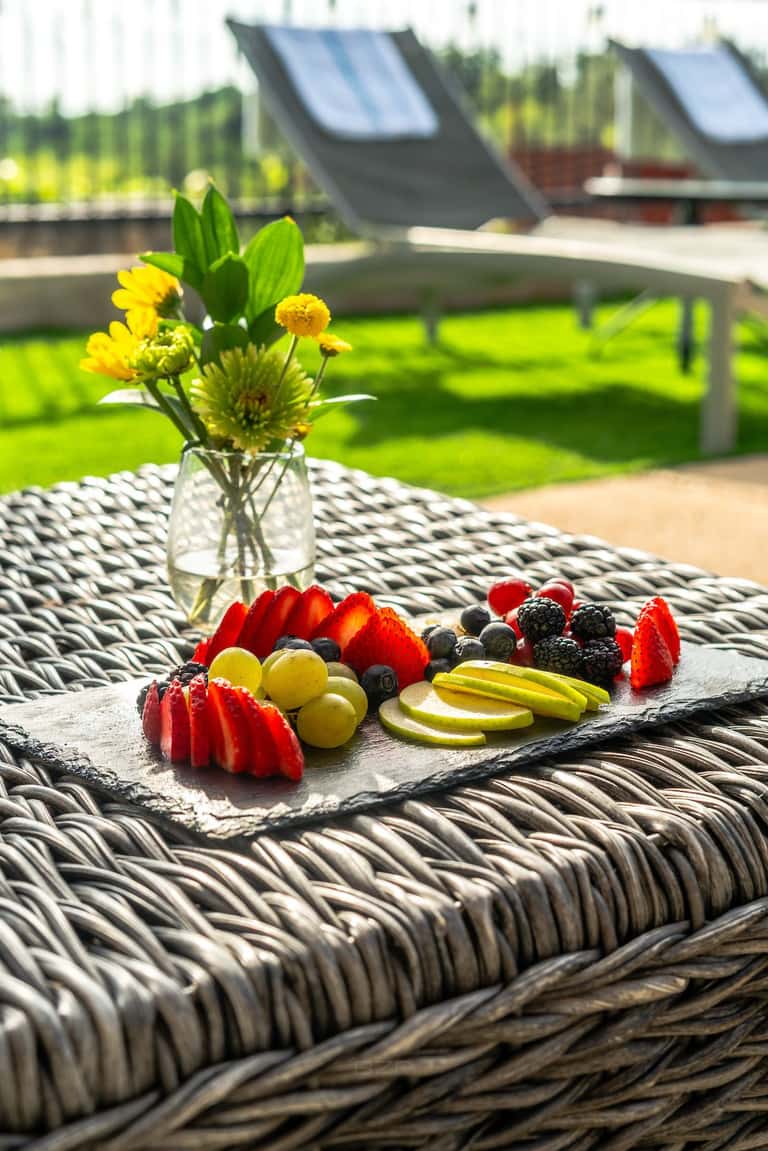Fresh fruit display on woven table with yellow flowers and lounge chairs visible in background