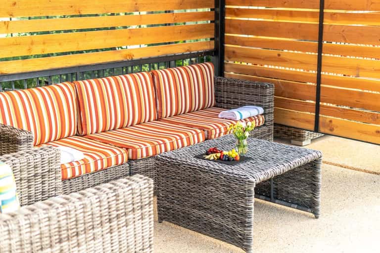 Outdoor patio with striped orange and white cushioned wicker seating, dark coffee table with flowers, and wooden privacy screens