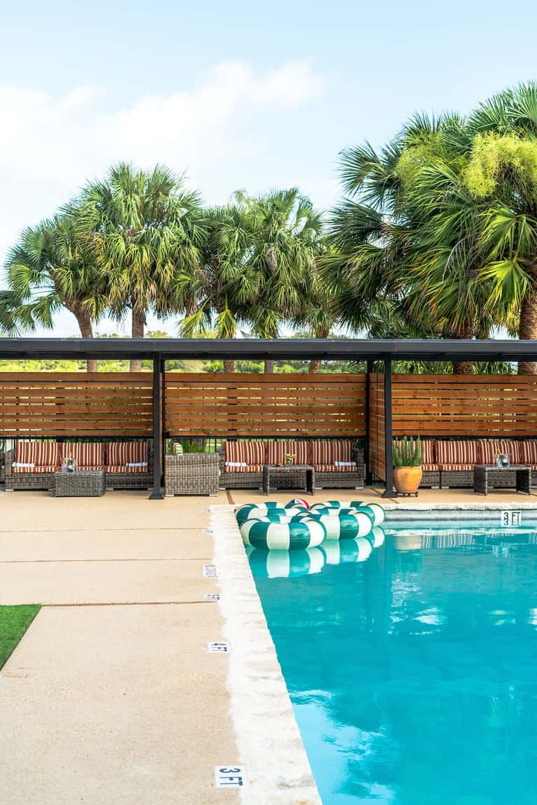 Resort pool deck with wooden privacy screens, wicker lounge chairs, green and white floats, and palm trees in background