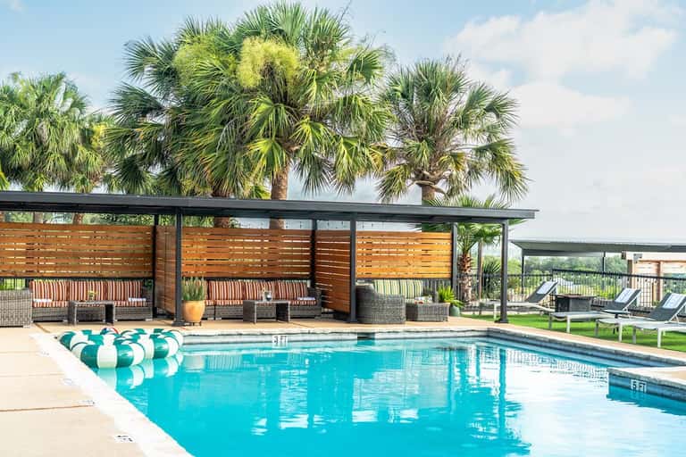 Expansive pool area with multiple wicker seating cabanas under wooden screens, palm trees, and green lounge chairs