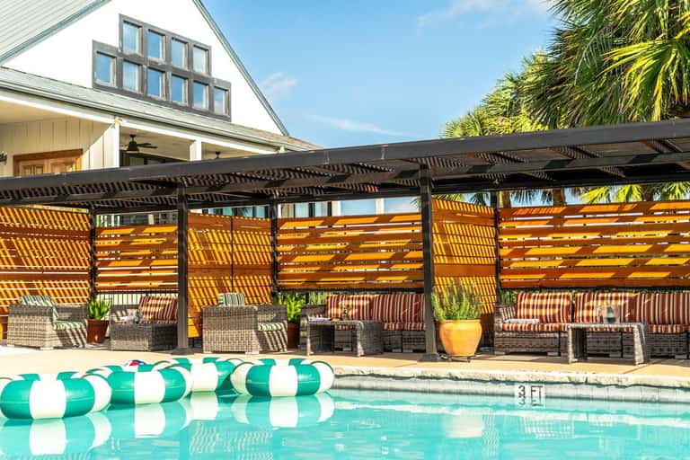 Luxury pool pavilion with angled wooden privacy screens, black metal pergola, striped seating, and white farmhouse architecture
