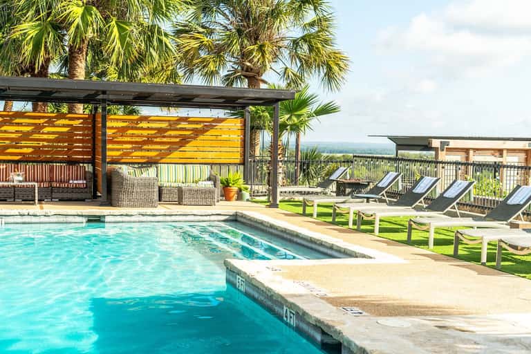 Resort pool with yellow pergola shade structure, palm trees, lounge chairs on grass, and distant ocean view