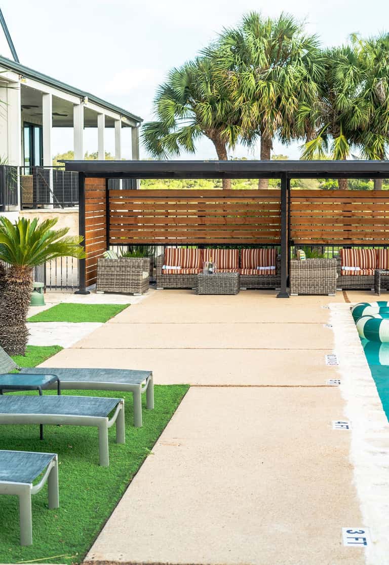 Wooden slat pergola structure over pool seating area with palm trees, lounge chairs, and concrete deck