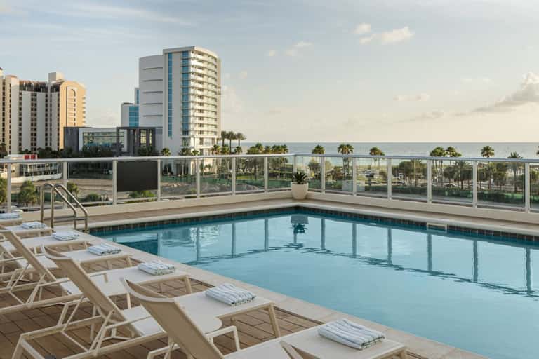 Beachfront hotel pool deck with lounge chairs, glass windscreen, ocean view, and palm trees under cloudy sky