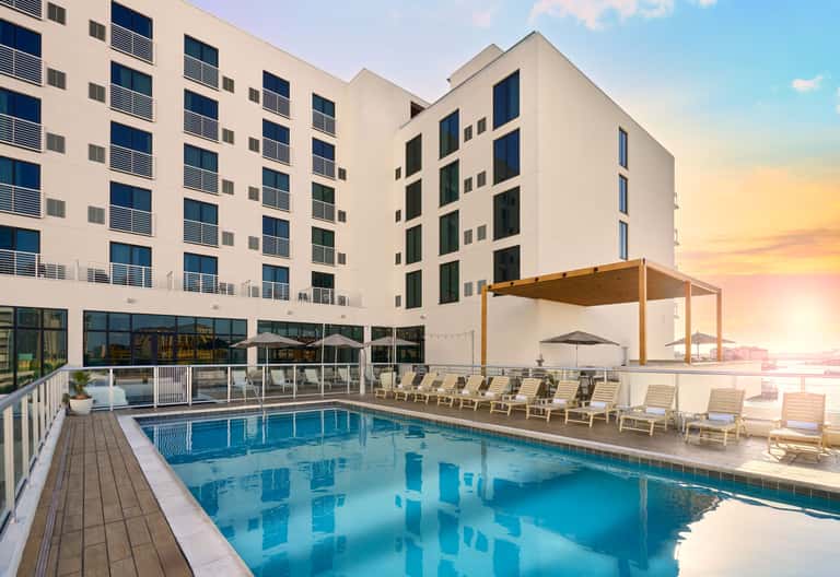 Resort pool at sunset with lounge chairs, pergola shade structure, and modern hotel building