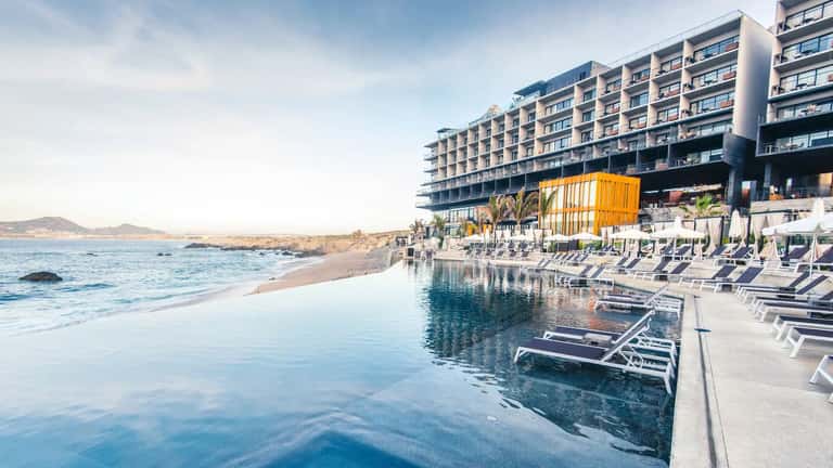 Beachfront resort with infinity pool, white loungers, and rocky coastline under blue sky