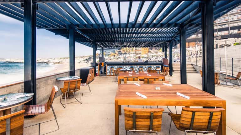Beachfront dining pavilion with wooden tables, orange chairs, and ocean views under pergola