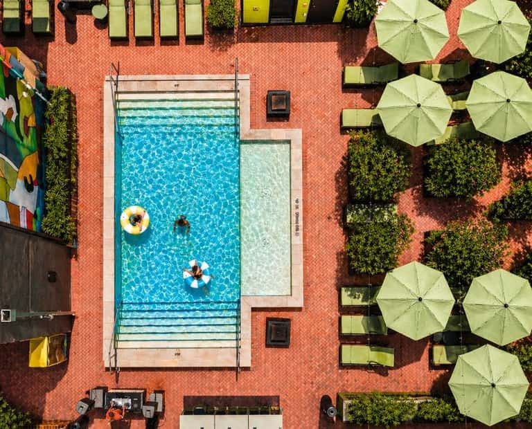 Aerial view of rectangular pool with blue water, two people swimming, yellow umbrellas, and terracotta brick patio with green plants