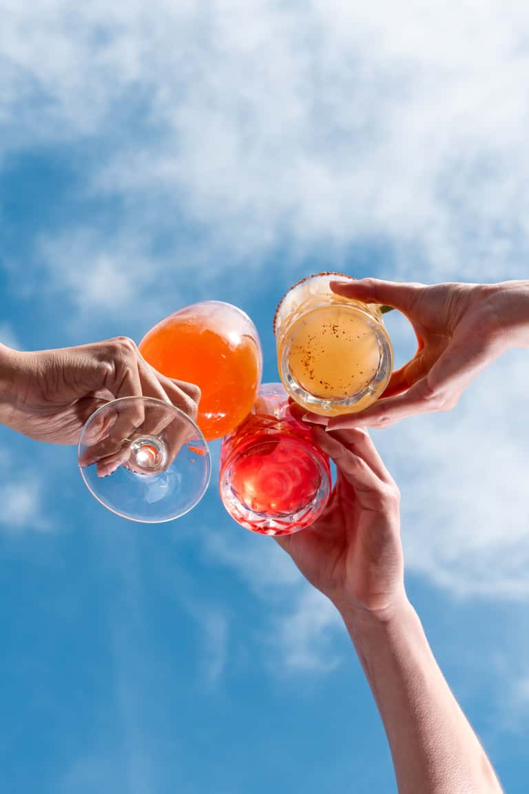 Four colorful cocktails held up against blue sky with white clouds