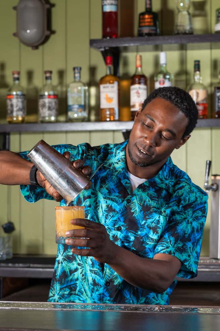 Bartender in tropical shirt mixing cocktail behind well-stocked bar