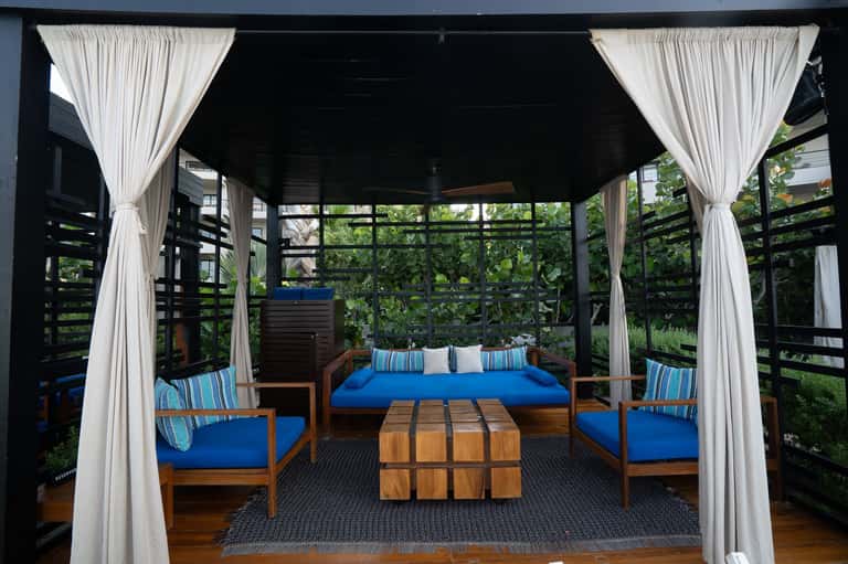 Modern outdoor lounge with blue seating, wooden furniture, white curtains, and garden views