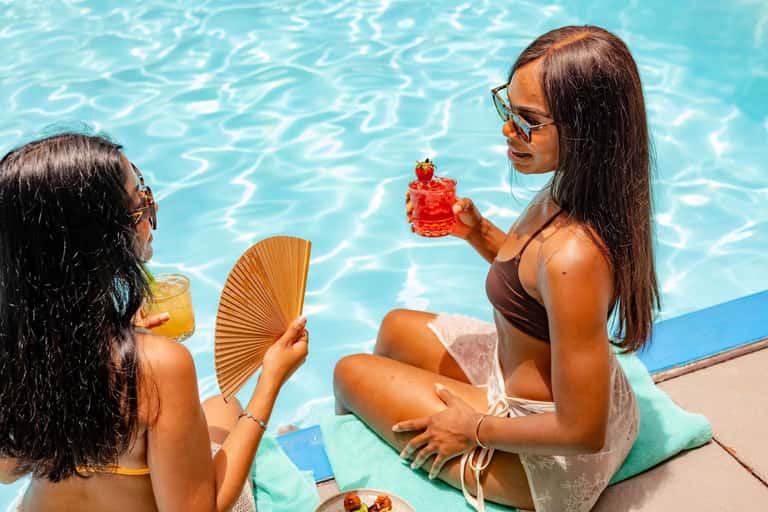 Two women by a swimming pool holding tropical drinks and a cocktail with fresh fruit
