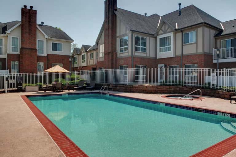 Resort outdoor swimming pool with turquoise water, surrounded by brick townhomes, white railing, lounge chairs, and umbrella