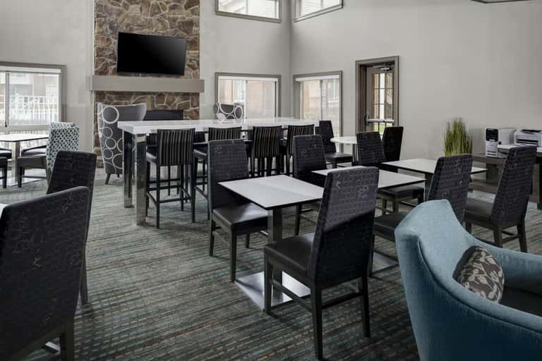 Modern hotel lounge with stone fireplace, TV, high-top dining tables, black chairs, and large windows overlooking exterior