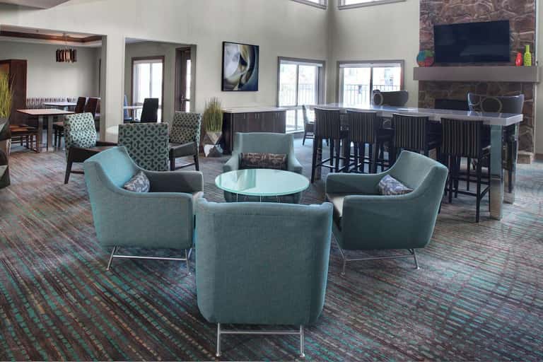 Contemporary hotel common area featuring turquoise armchairs, round glass table, stone fireplace with TV, bar seating, and natural light