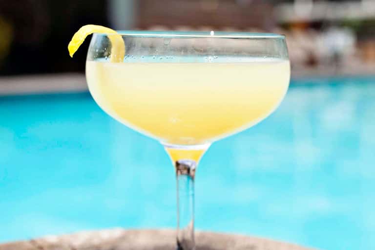 Pale yellow cocktail with lemon twist garnish served poolside with turquoise pool water in background