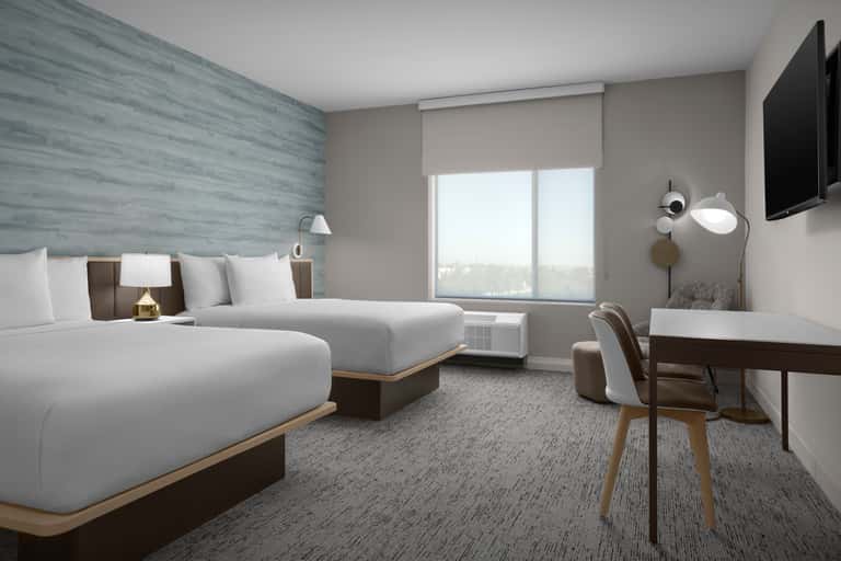 Modern hotel room with two queen beds, blue accent wall, desk, TV, and large window with city views