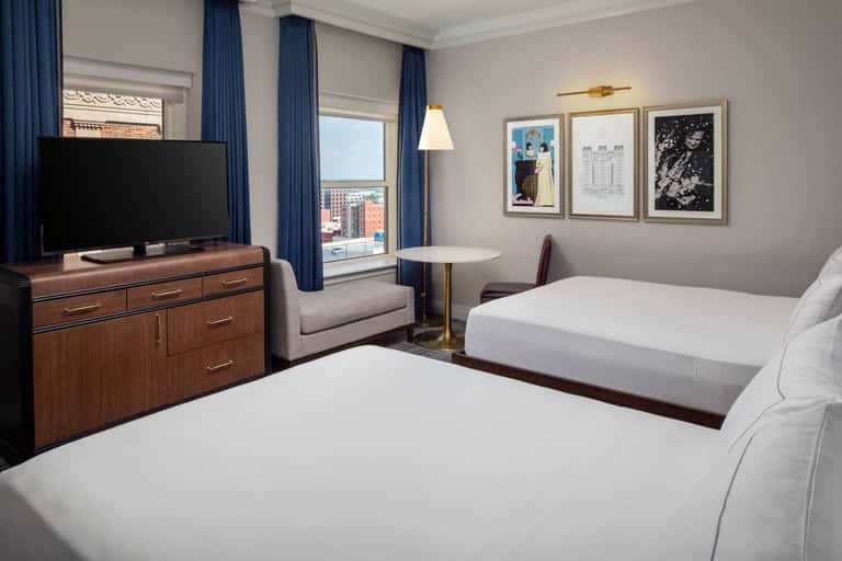 Hotel guest room with two beds, wood dresser, TV, desk area, and city views through large windows